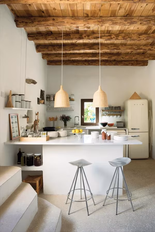 Cocina blanca de la casa de Nani Marquina en Ibiza con vigas de sabina, lámparas de mimbre y taburetes