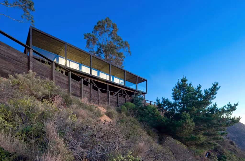 Casa Till vista desde abajo, una franja horizontal de madera oculta en la ladera bajo el cielo azul