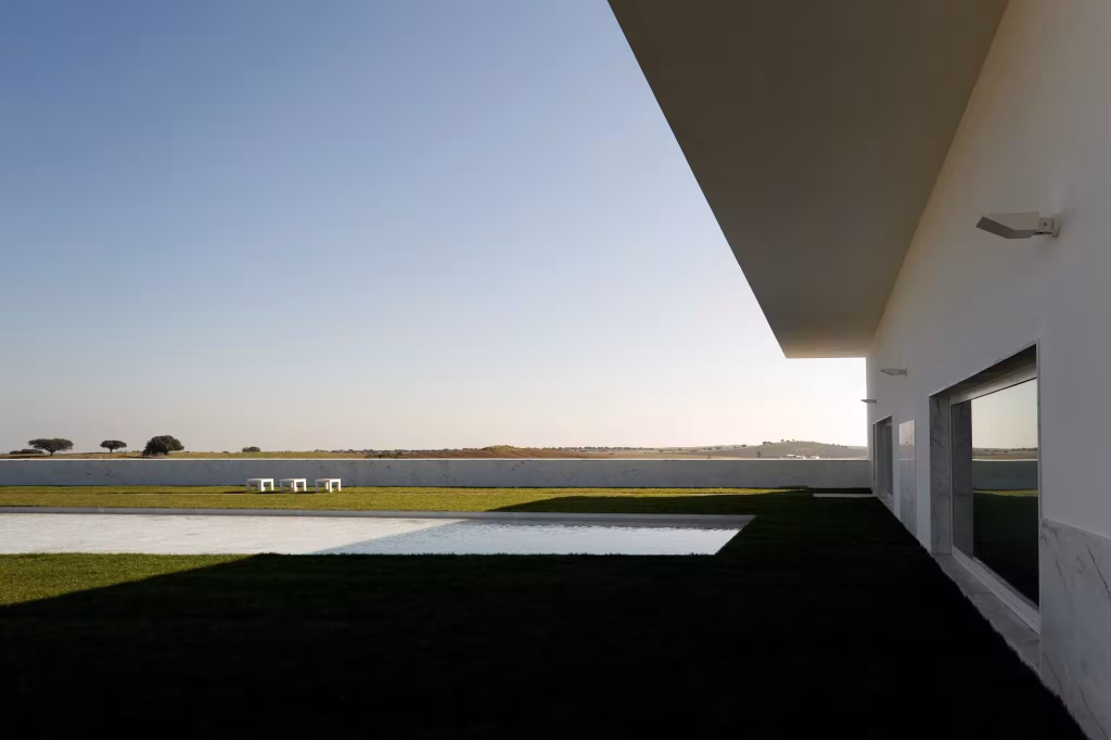 Fachada blanca y voladizo de la Adega Maior al atardecer, con césped y lámina de agua frente a la llanura