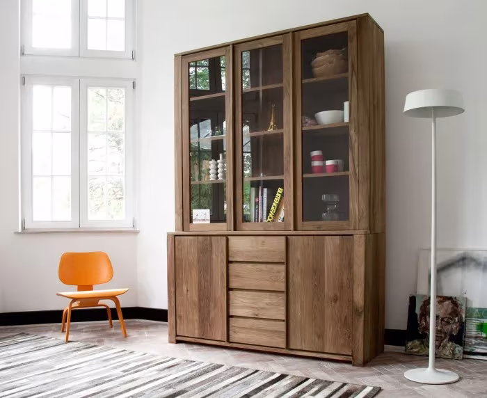 Vitrina alta de madera maciza de Ethnicraft con puertas de vidrio y cajones, junto a una silla naranja