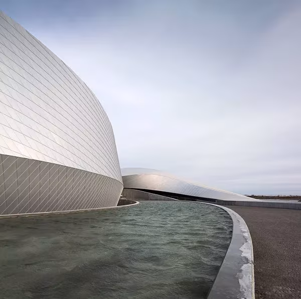 Muros curvos plateados de El Planeta Azul de 3XN que se abren en torno a un estanque bajo cielo gris