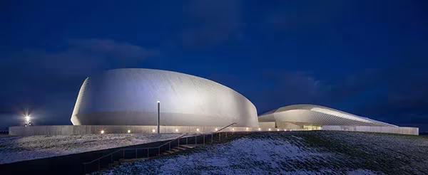 Vista nocturna del acuario El Planeta Azul de 3XN con su cubierta plateada iluminada en Copenhague