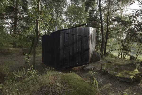 Cabaña de madera quemada negra de Uhlik Architekti cerrada entre árboles y rocas del bosque bohemio