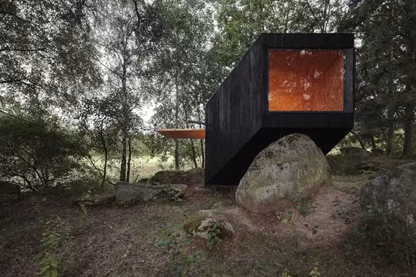 Extremo de la cabaña negra apoyado sobre una gran roca con su ventanal iluminado en Bohemia