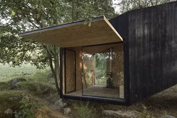 Cabaña negra con su gran ventana abatible abierta como alero sobre el claro del bosque