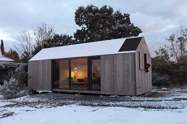 Casa Eficiente ÁPH80 de ÁBATON con revestimiento de madera, placa solar y gran ventanal sobre la nieve