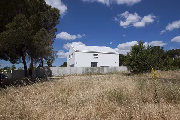 Casa Eficiente de ÁBATON blanca entre pinos, vista desde un prado de hierba seca bajo un cielo azul con nubes