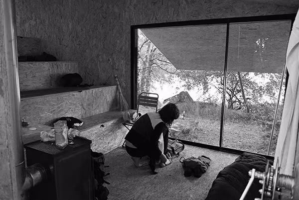 Persona agachada en el interior de la Cabaña en Bohemia junto a la estufa y el ventanal, en blanco y negro