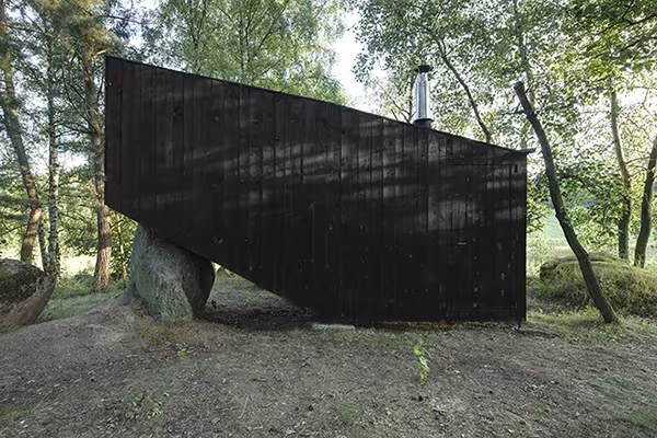Volumen de madera negra de la Cabaña en Bohemia apoyado en grandes rocas, con chimenea metálica