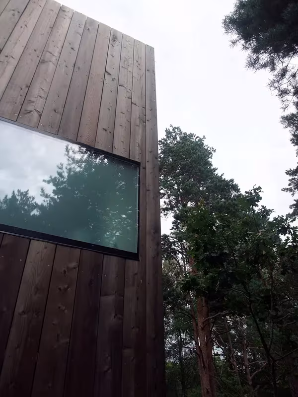 Esquina de la Casa Cubo en Veierland revestida de tablas de madera oscura con una ventana alargada entre pinos