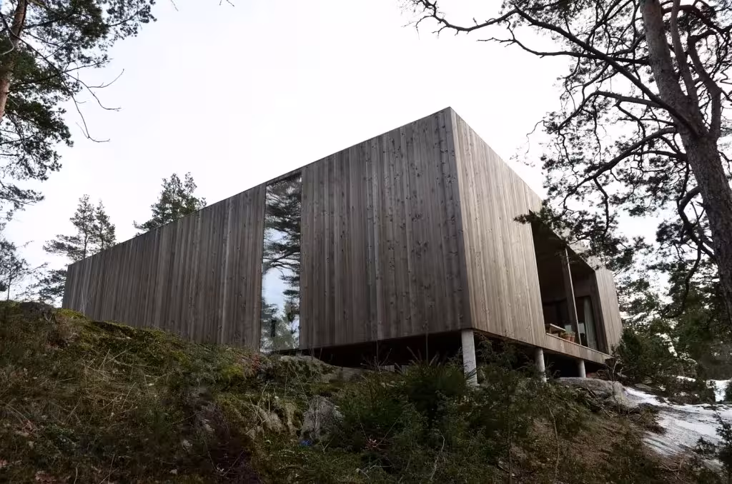 Casa Cubo en Veierland vista desde abajo, un prisma de madera grisácea apoyado sobre pilares en la roca