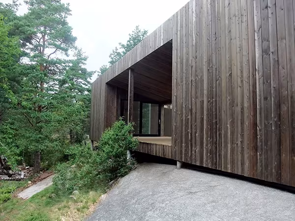 Casa Cubo en Veierland con fachada de madera oscura y porche abierto sobre un camino de grava entre pinos