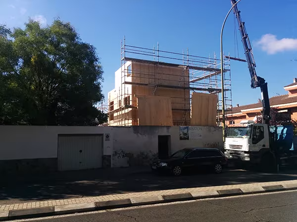 Grúa montando los paneles de madera maciza de una Casa Eficiente de ÁBATON tras un muro de parcela