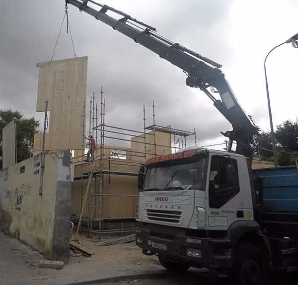 Camión grúa izando un panel de madera maciza para una Casa Eficiente de ÁBATON entre andamios