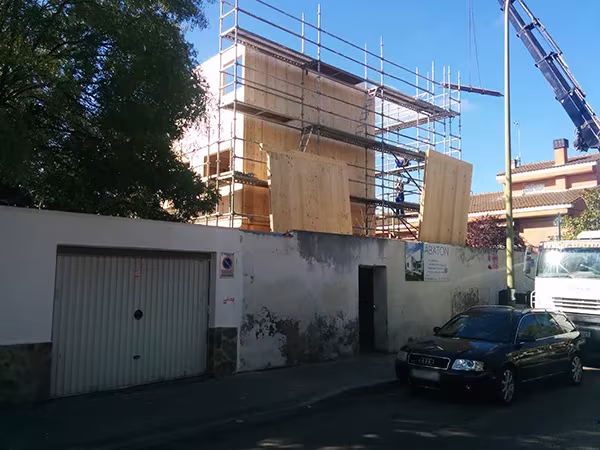 Estructura de paneles de madera de una Casa Eficiente de ÁBATON elevándose sobre la tapia de la calle