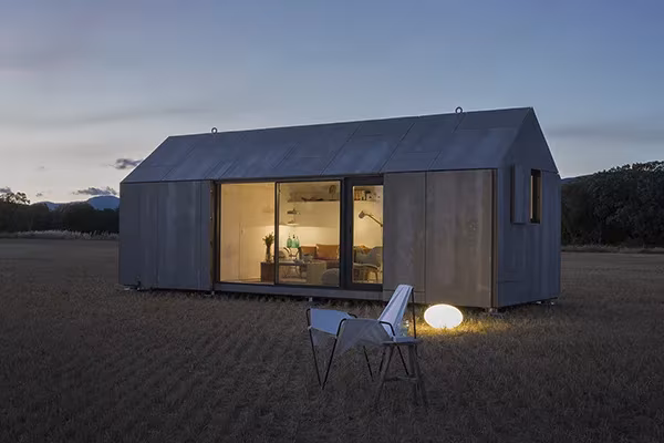 Casa ÁPH80 de ÁBATON al anochecer con el interior iluminado tras el ventanal y una lámpara en la hierba
