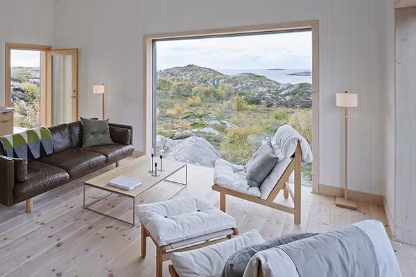 Salón de la Vega Cottage con sofá de cuero y butacas ante un gran ventanal abierto al paisaje costero