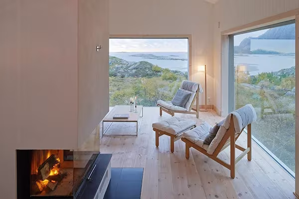 Salón de la Vega Cottage con chimenea, butacas de madera y ventanales hacia el fiordo y las islas