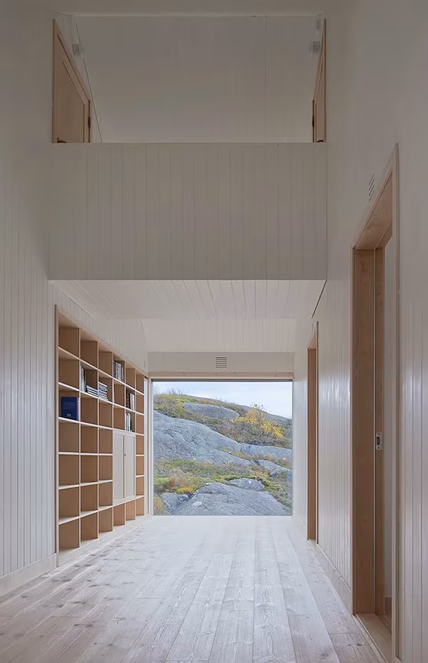 Pasillo blanco de la Vega Cottage con estantería de madera que termina en una ventana hacia las rocas