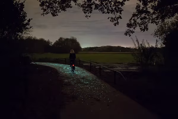 Ciclista lejano en el carril bici luminoso de Daan Roosegaarde junto a una valla y campos al crepúsculo