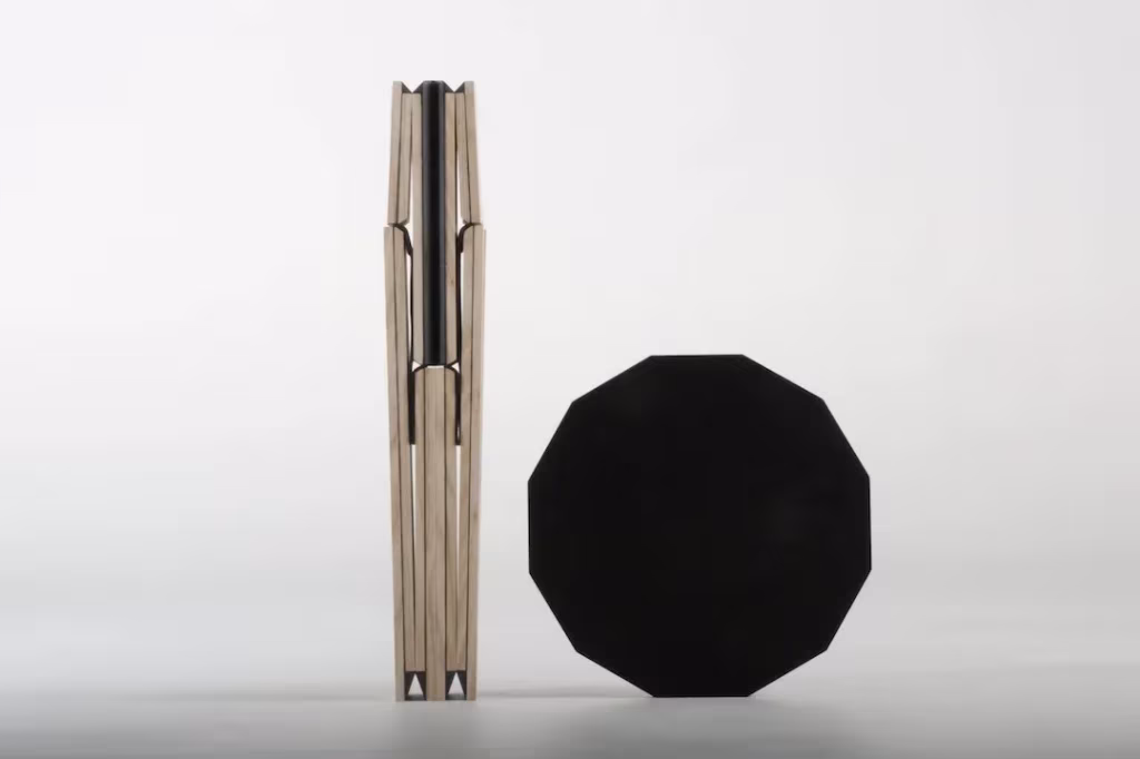 Taburete plegado de patas de madera junto a su asiento negro poligonal apoyado de canto sobre fondo blanco