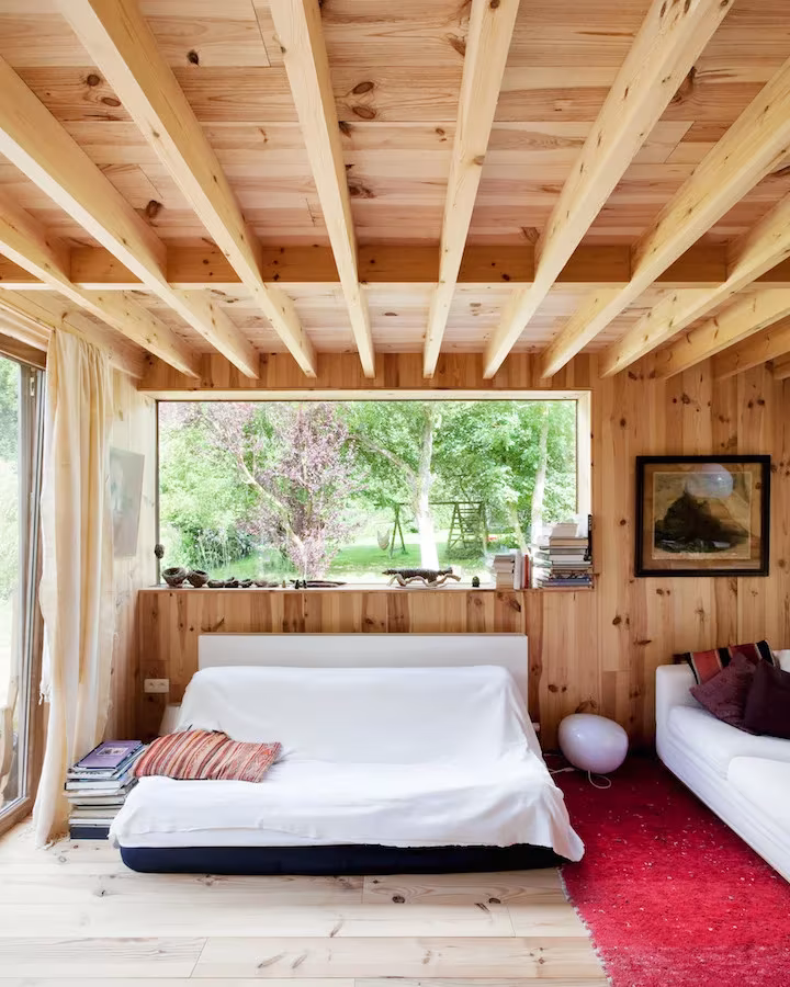 Dormitorio con vigas de pino vistas y cama blanca bajo un ventanal al jardín en la Casa de las 14 fachadas
