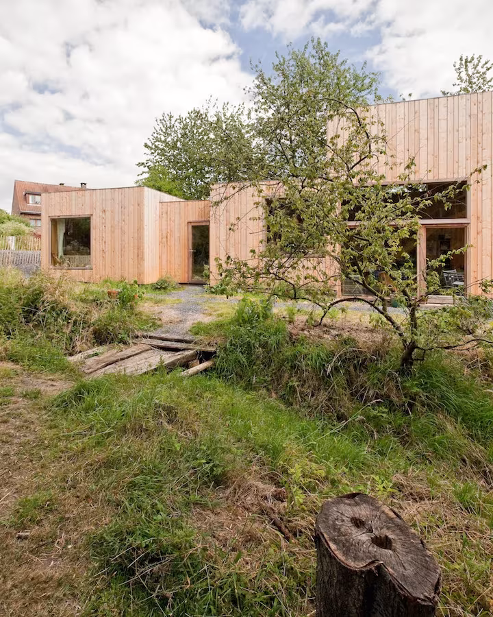 Volúmenes de madera de la Casa de las 14 fachadas entre hierba y árboles frutales en un terreno irregular