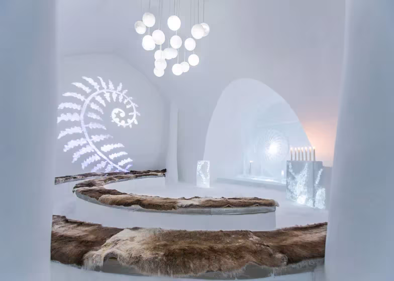 Suite del Icehotel con espiral luminosa tallada en la pared, lámpara de esferas y cama redonda con pieles