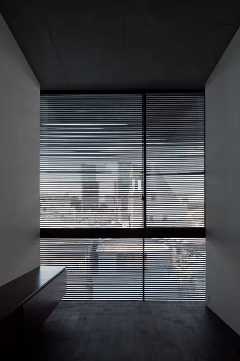 Estancia en penumbra de la Casa Lattice con un ventanal tamizado por densas lamas de madera