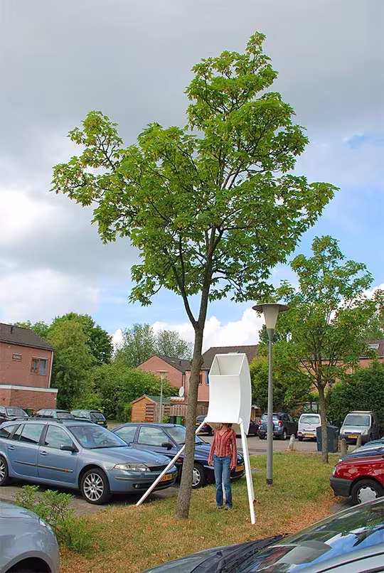 Persona mirando dentro de un visor blanco de Rob Sweere apoyado en un árbol junto a un aparcamiento