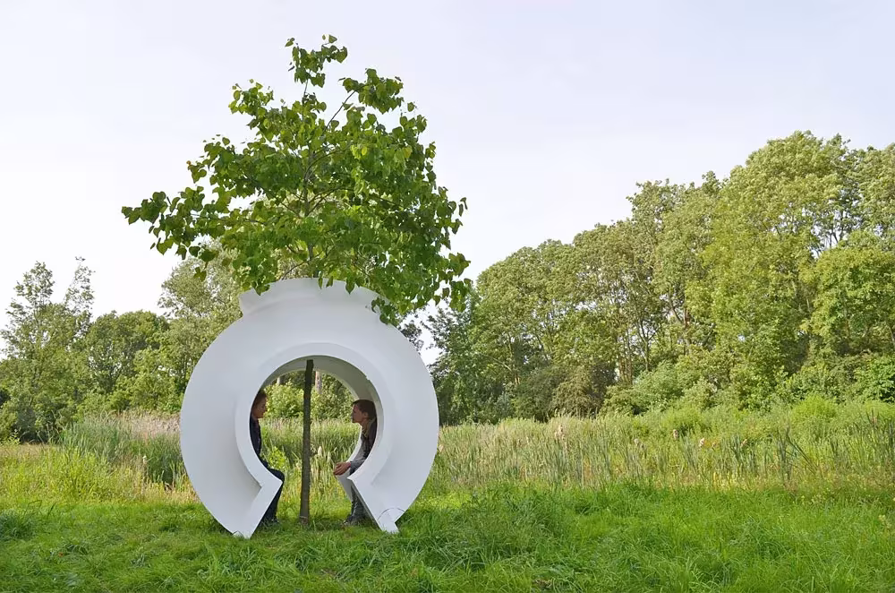 Love Tree de Rob Sweere, un anillo blanco que abraza el tronco de un árbol joven en un prado verde