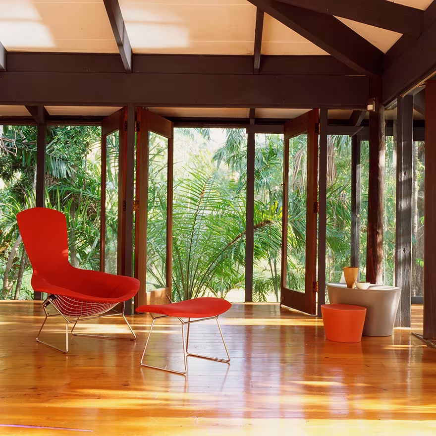 Butaca y reposapiés Bird de Harry Bertoia en rojo en un porche de madera abierto a una vegetación tropical
