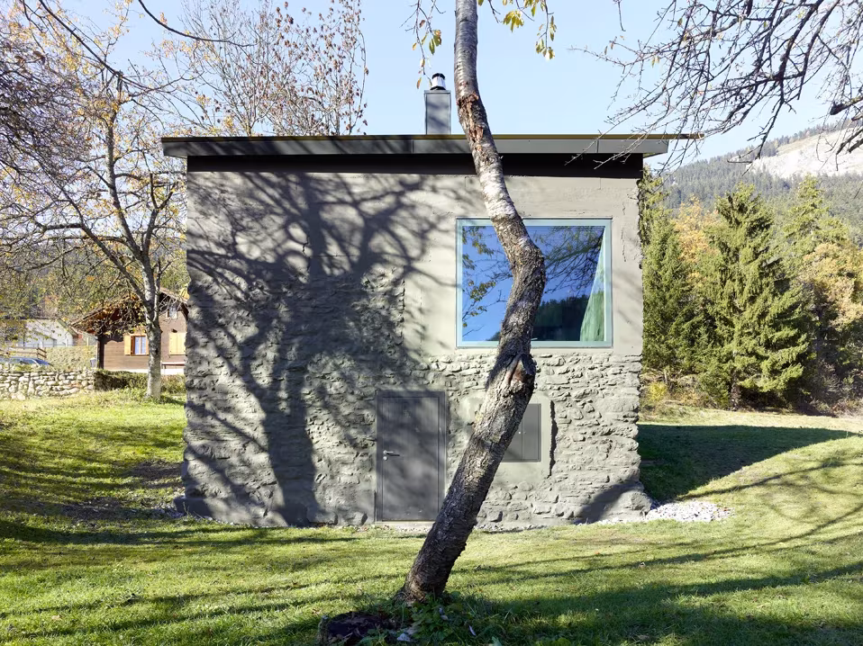 Mayen de piedra rehabilitado por Savioz Fabrizzi en un prado con árboles y una ventana enmarcada en metal