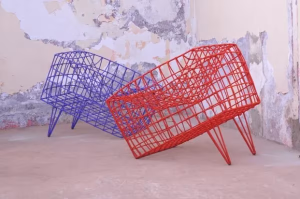 Dos sillones de varilla metálica, uno azul y otro rojo, de formas facetadas ante un muro desconchado