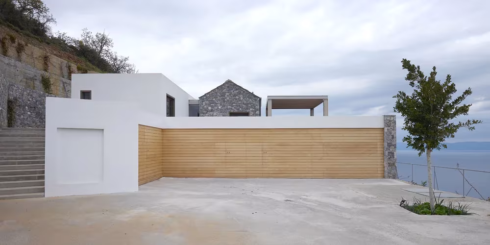 Villa Melana en Grecia, volúmenes blancos y de piedra con muro de madera junto a una explanada de hormigón