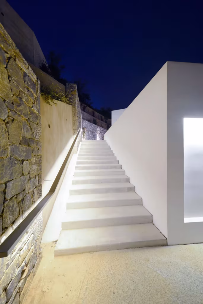 Escalera blanca iluminada de noche entre un muro de piedra y un volumen encalado en Villa Melana