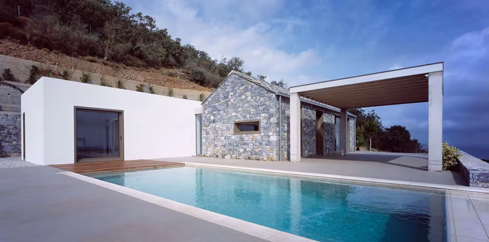 Piscina de Villa Melana a pleno sol con el volumen blanco, la casa de piedra y la pérgola al fondo