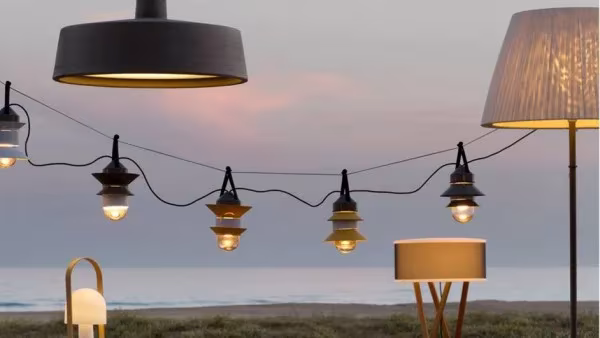 Lámparas de Marset en una terraza frente al mar al atardecer, con guirnalda de luces y lámpara de pie