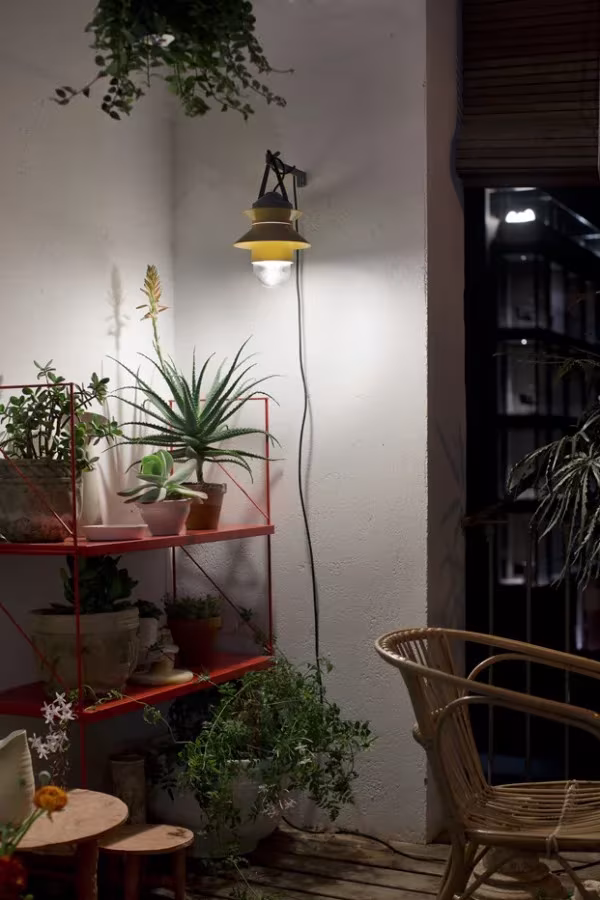 Rincón de patio con aplique Marset iluminado, estantería roja con plantas y butaca de mimbre