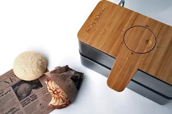 Caja panera de bambú y metal negro junto a un panecillo y papel de periódico