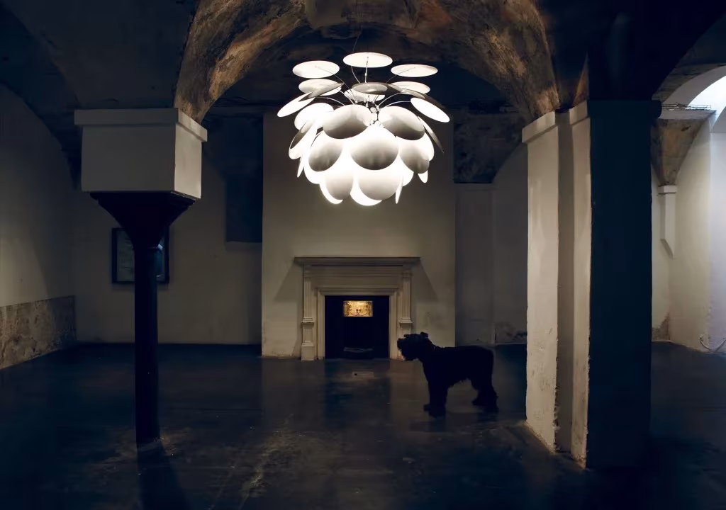Lámpara Discocó gigante de Marset colgada en una sala abovedada en penumbra, con un perro junto a la chimenea