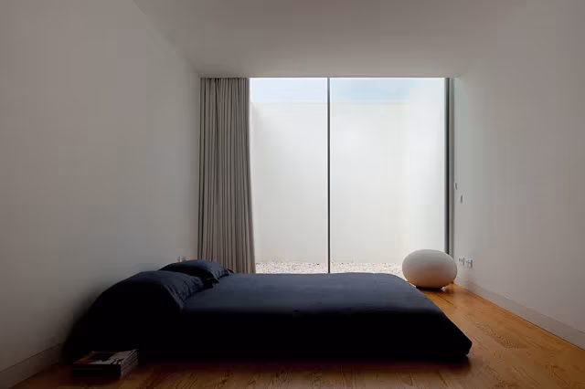Dormitorio minimalista de la Casa Leiria con cama baja oscura y luz cenital por un patio