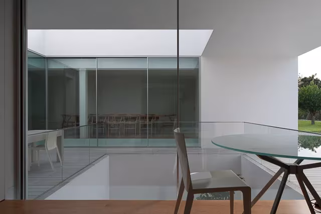 Comedor de la Casa Leiria con mesa de cristal junto a un hueco a doble altura y paredes blancas