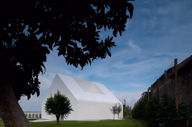 Casa Leiria vista entre ramas oscuras, con su silueta blanca a dos aguas y un árbol delante