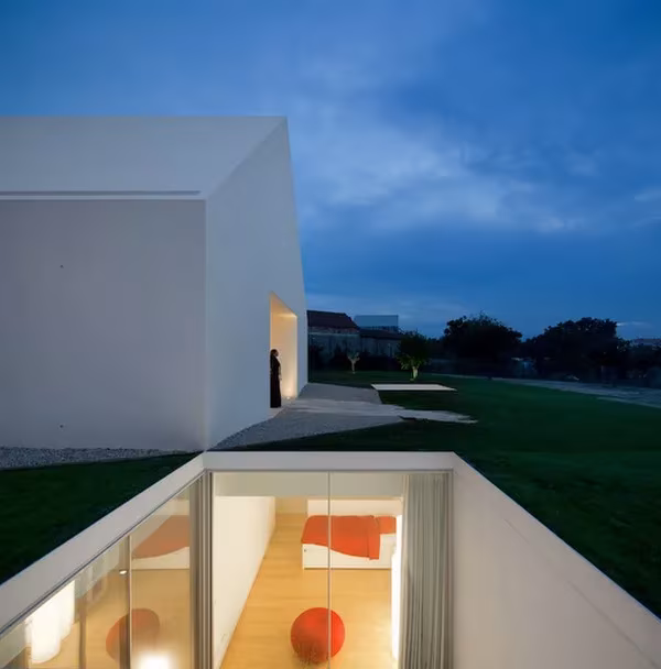 Patio hundido de la Casa Leiria iluminado de noche, con interior de muebles rojos bajo el volumen blanco