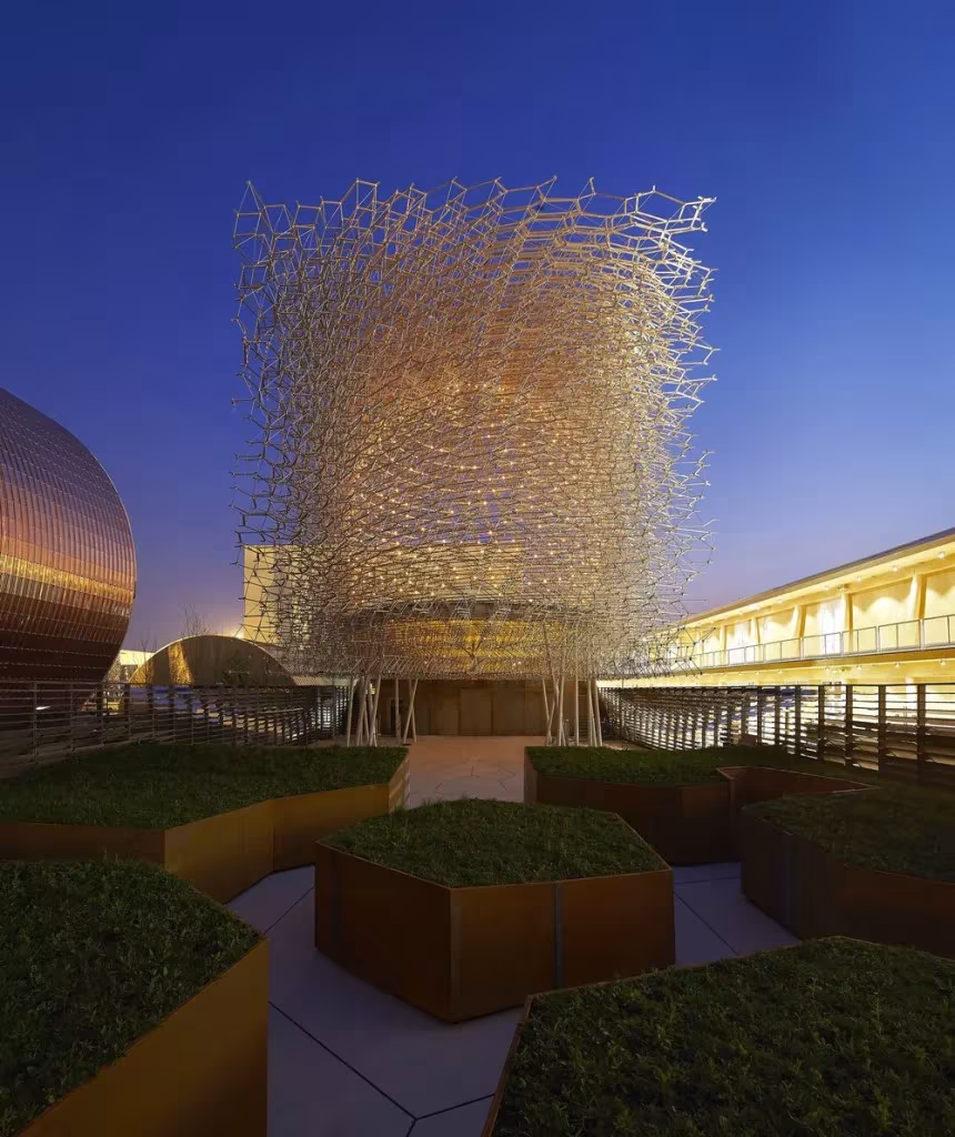 La Colmena iluminada al anochecer sobre jardineras de acero corten con vegetación en el pabellón de Reino Unido de la Expo de Milán