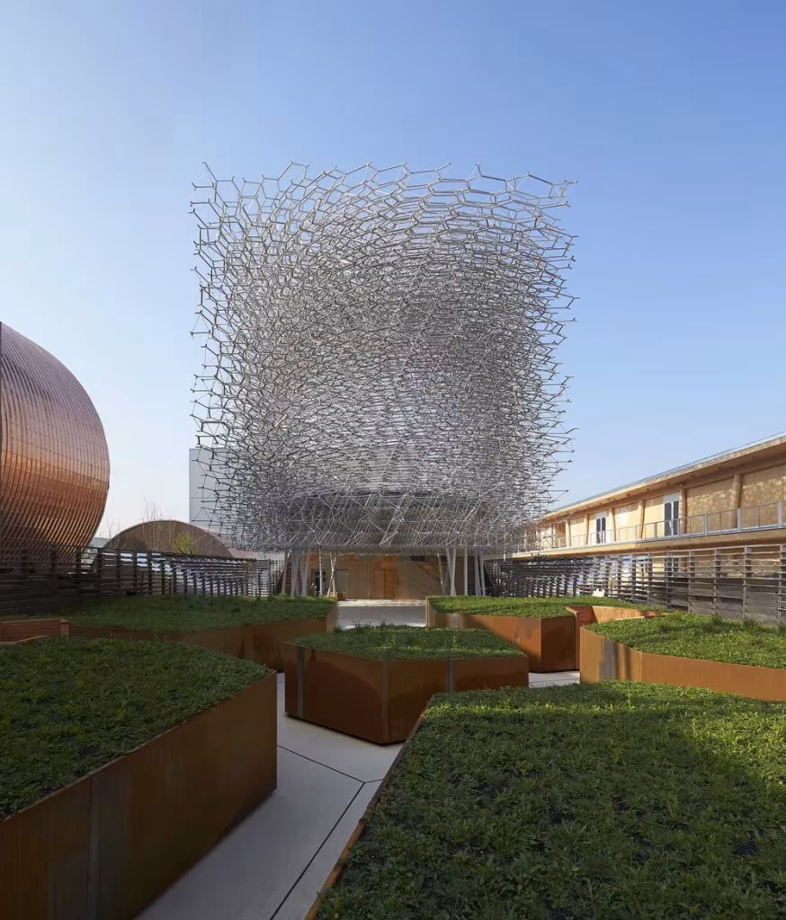 Cubo de malla metálica de La Colmena con luz de día, rodeado de jardineras de acero corten y prados en la Expo de Milán