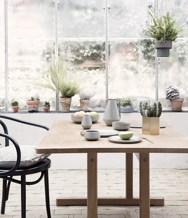 Mesa de madera con vajilla gris de Ferm Living, silla curvada y macetas con plantas junto a un gran ventanal
