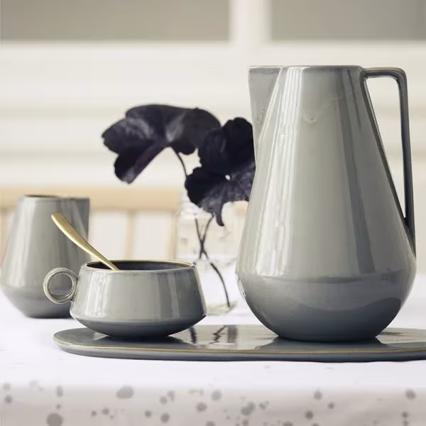 Jarra y tazas de cerámica gris esmaltada de Ferm Living sobre una bandeja, con flores oscuras en un jarrón
