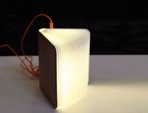 Lámpara Lumio entreabierta con tapas de madera y cable naranja, emitiendo luz blanca sobre una mesa